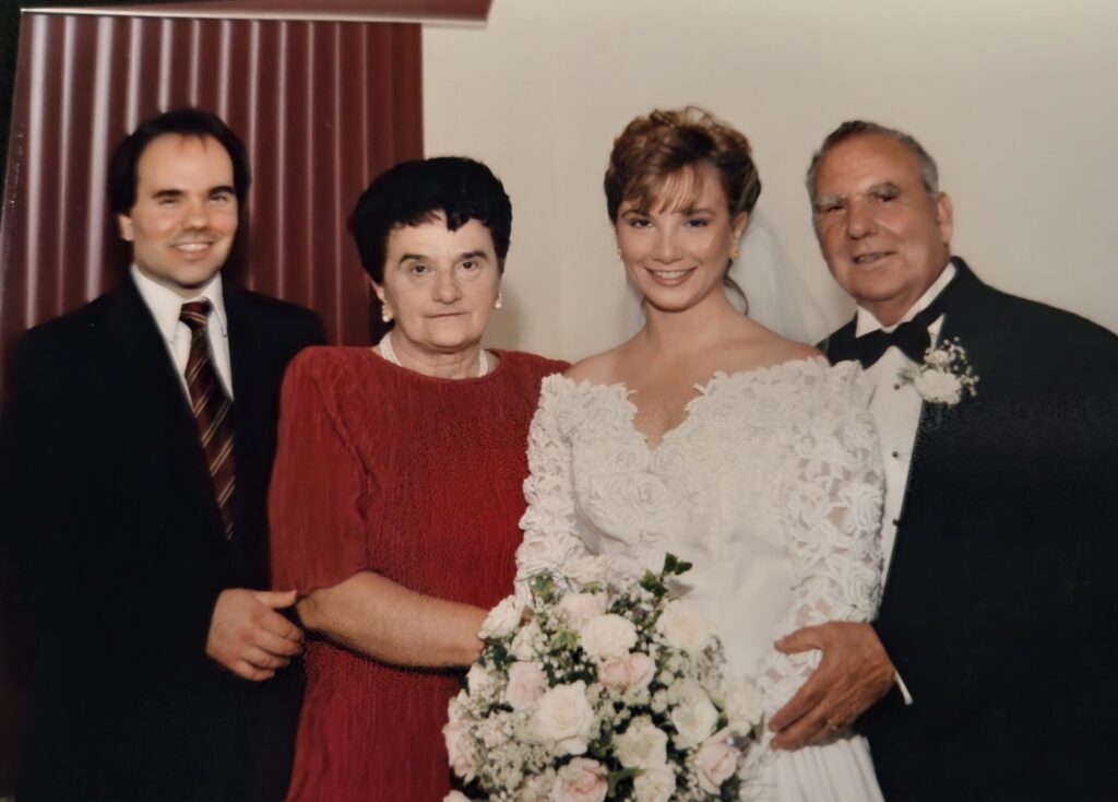 A color photograph with a young White bride standing in the middle of an elderly White man in a tuxedo, an elderly White woman in a dress, and a young White man in a suit and tie. 