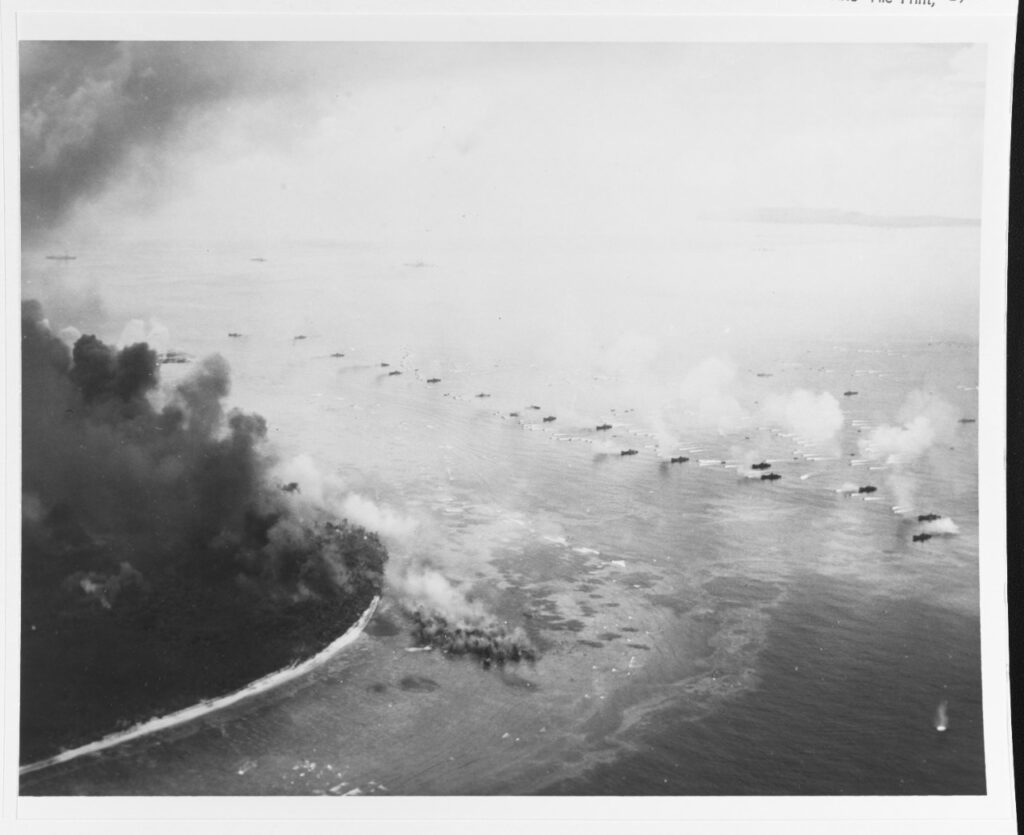 A black and white aerial image of several landing vessels off the shore of an island.