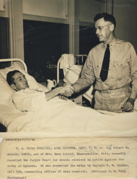 A typed declaration of the award. A black and white image of a young White man in a hospital bed shaking hands with another, older White man in uniform.