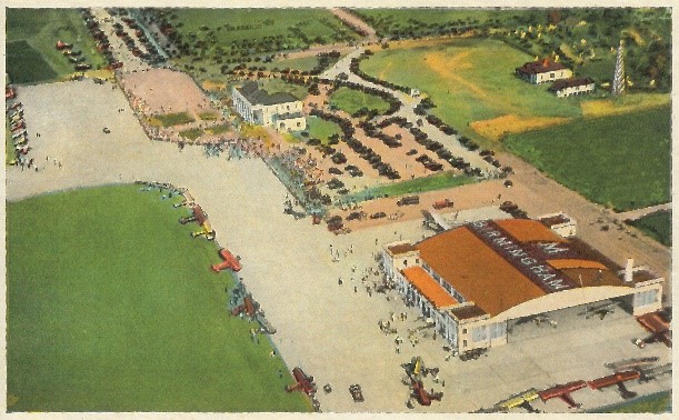 A color postcard showing an aerial view of the airport. There are several planes parked on the runway. The top of the airplane hangar reads “Birmingham.”