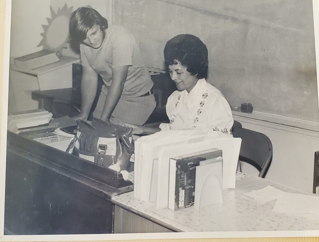 A black and white image of an older woman sitting at a desk talking with a student.