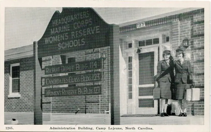 A black and white postcard image of two women in uniform standing outside a brick building. In front is a sign reading, “Headquarters Marine Corps Women’s Reserve Schools.”