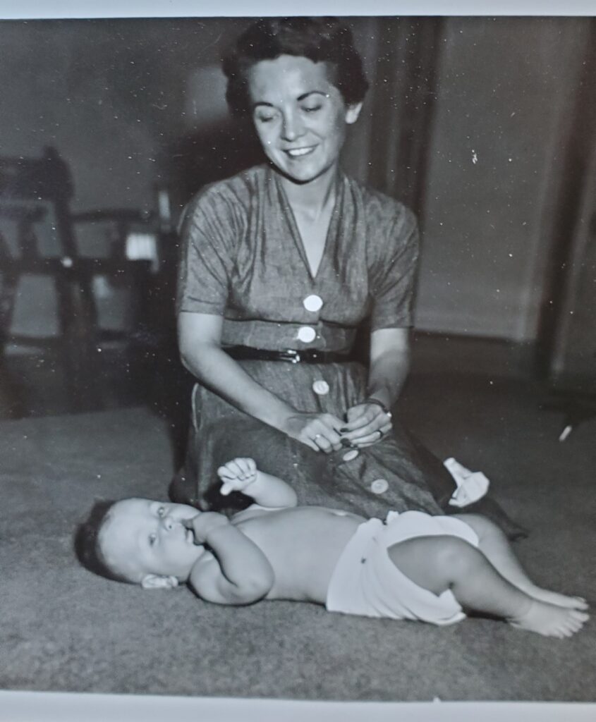 A black and white image of a young White woman kneeling next to a baby on the living room floor.