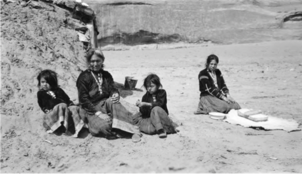 A black and white image of an older Native American woman sitting on the ground with two small children and a young woman. 