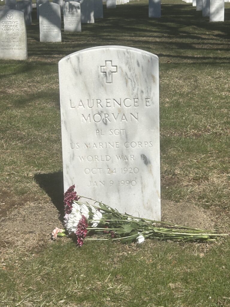 A color image of an arched white marble headstone engraved with a cross at the top and “Laurence E. Morvan PL SGT US Marine Corps World War II Oct 24 1920 Jan 9 1990.”
