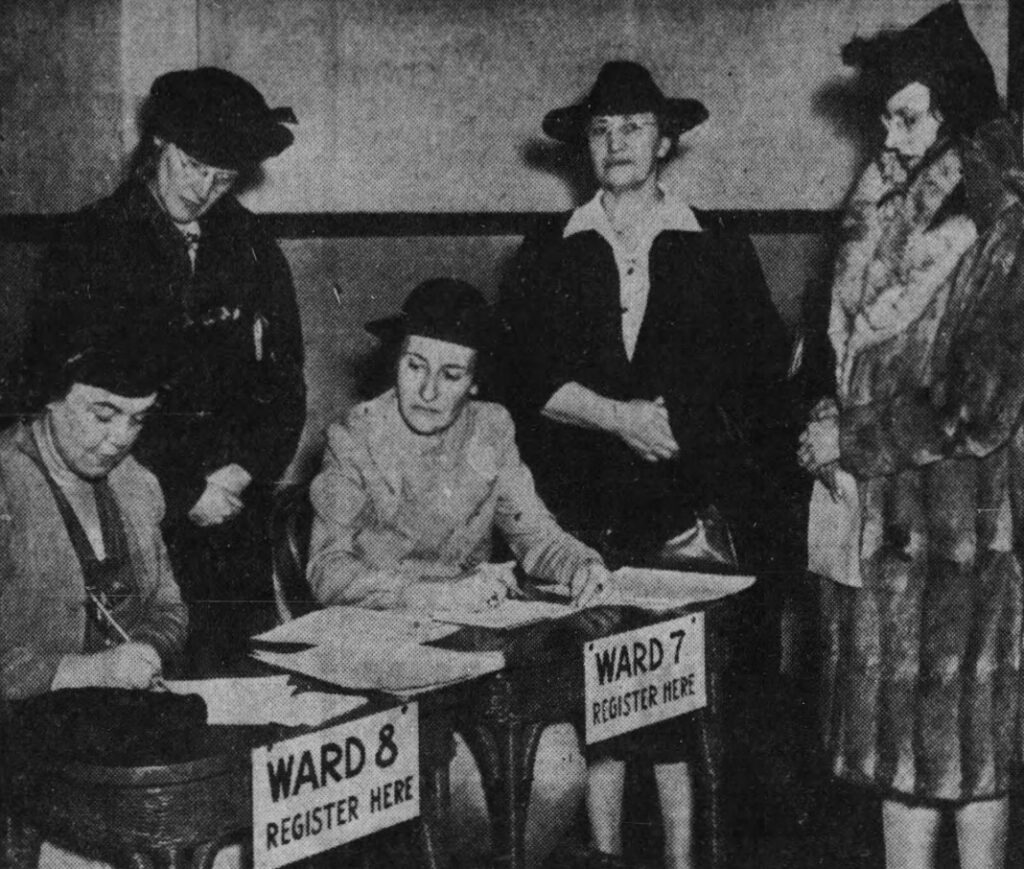 A black and white image of two women sitting behind desks signing people up that are in line.