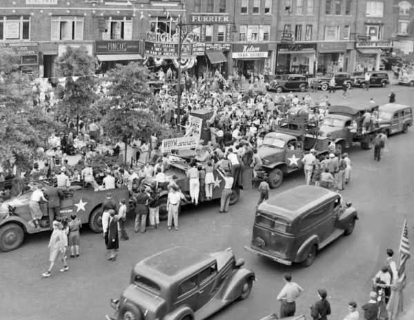 A black and white aerial image of several cars parked in a city square with dozens of people standing around.