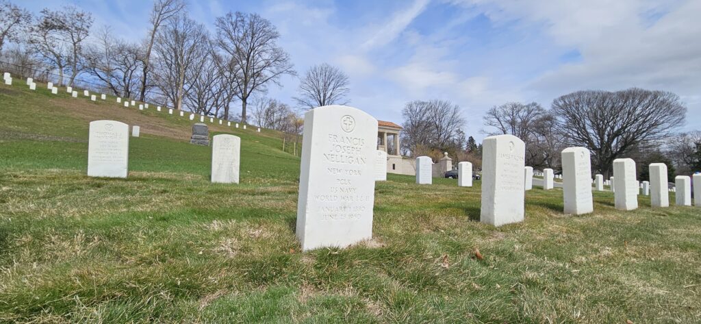 A color photograph of an arched white marble headstone engraved with a cross and “Francis Joseph Nelligan New York PCLK US Navy World War I & II January 1 1880 June 28 1950.”