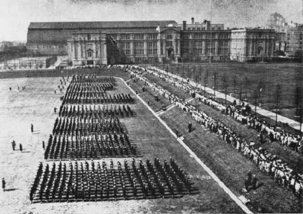 A black and white aerial image of dozens of women in uniform standing in formation, with dozens more watching.