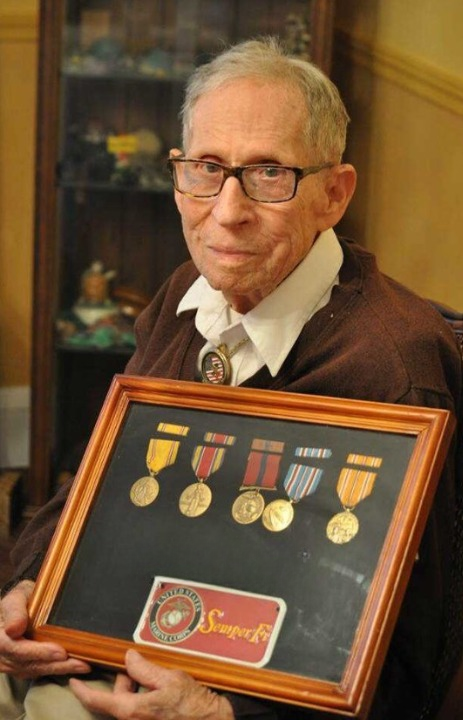 A color image of an elderly White man dressed up and holding a framed shadowbox of medals.