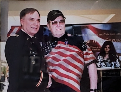 A color image of an older White man in uniform shaking hands with an elderly White man in an American flag t-shirt. 