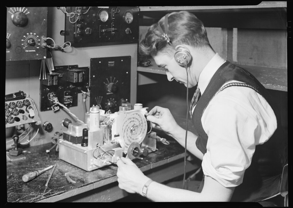 A black and white image of a young White man sitting at a desk working on a radio.