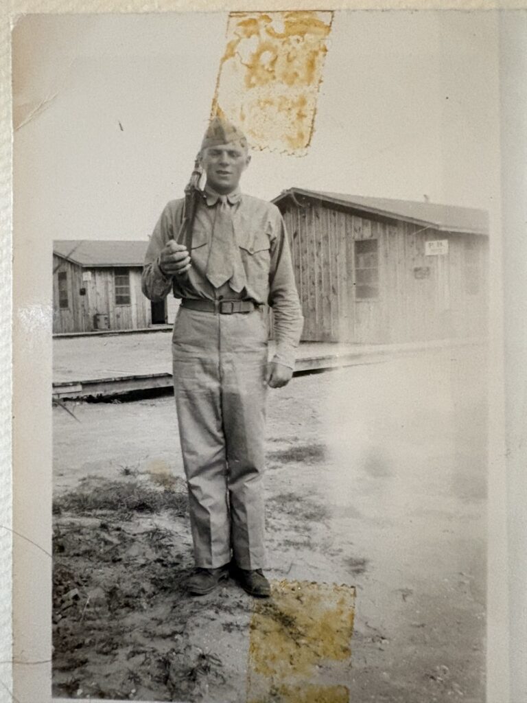 A black and white photograph of a young White man in a uniform holding a gun outside some barracks.