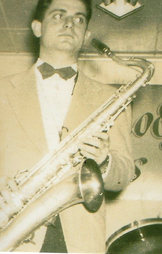 A black and white image of a young White man in a suit and tie holding a saxophone.