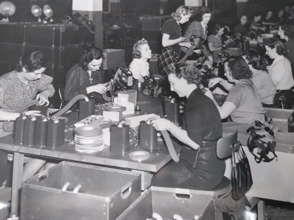 A black and white photo of several women sitting at a large table assembling gas masks.